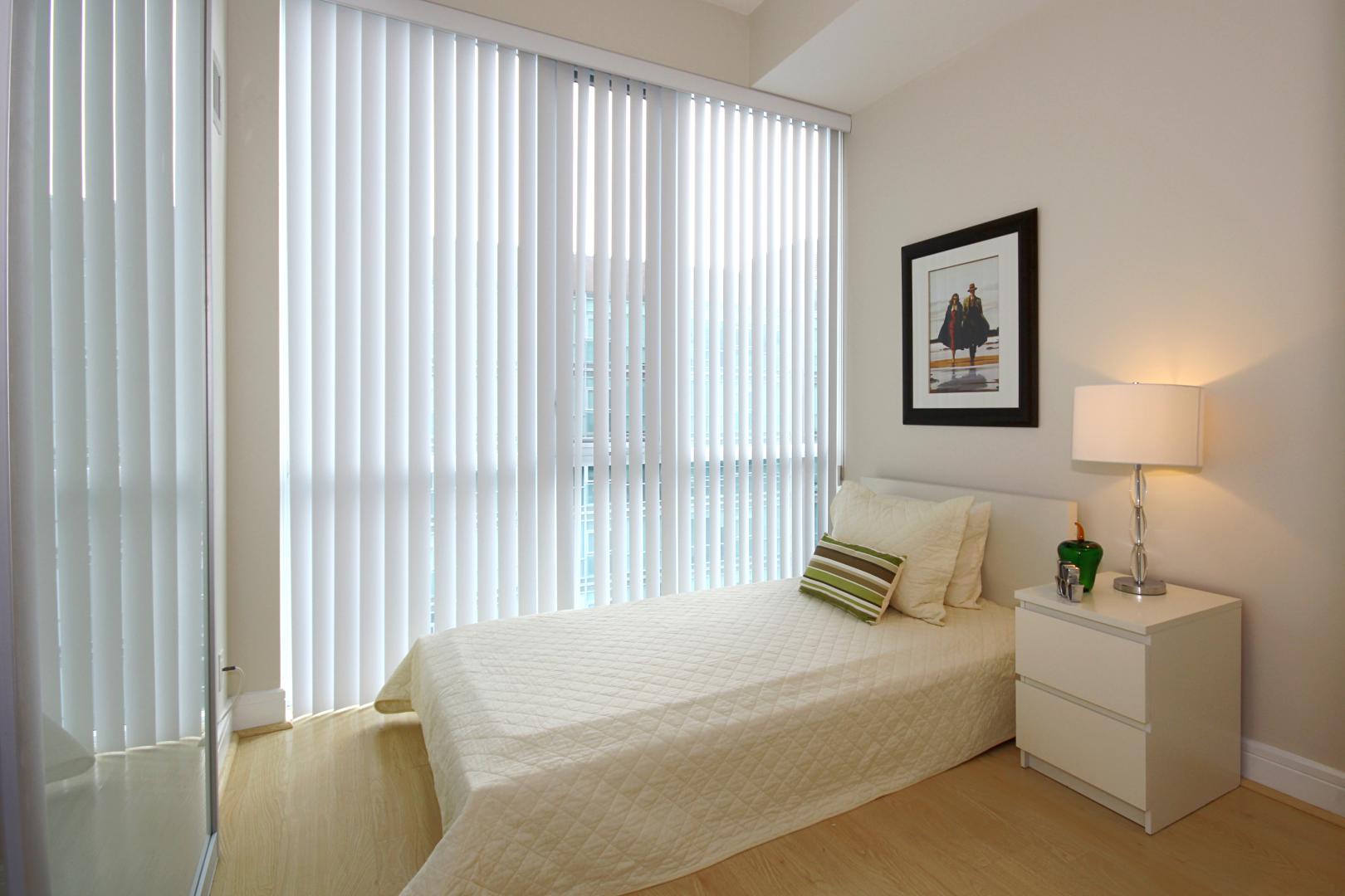 A bedroom with white vertical blinds on a large window.
