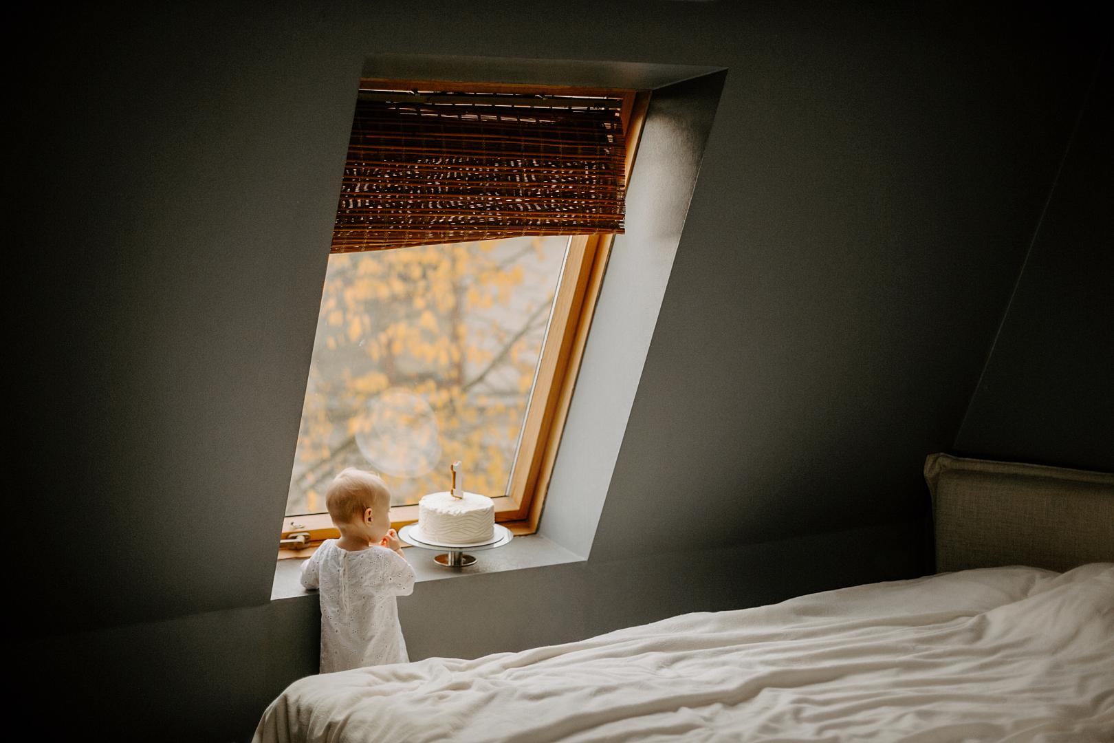 A young child standing next to an attic window with inside-mount shades.
