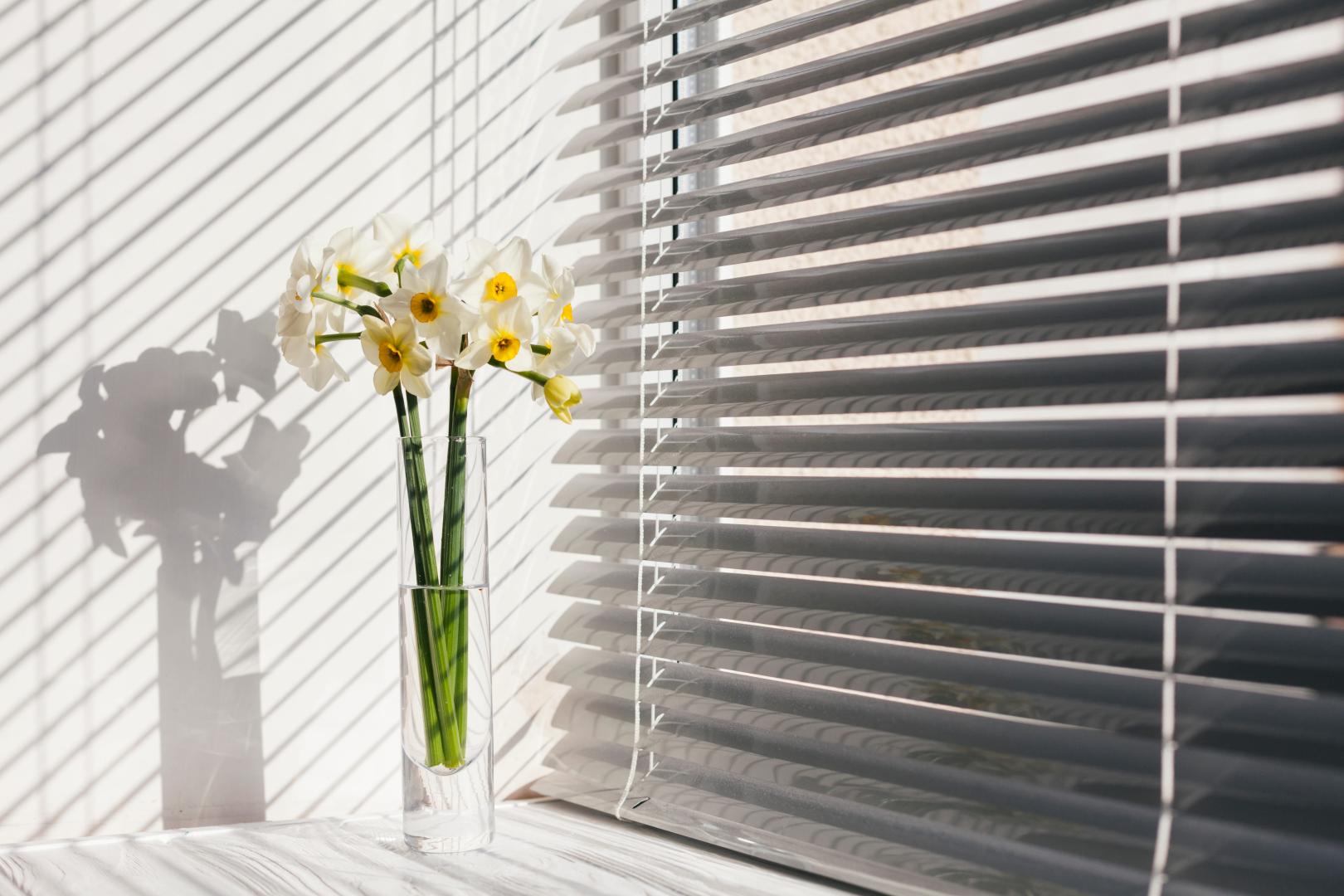 A window with white blinds and flowers in a vase.