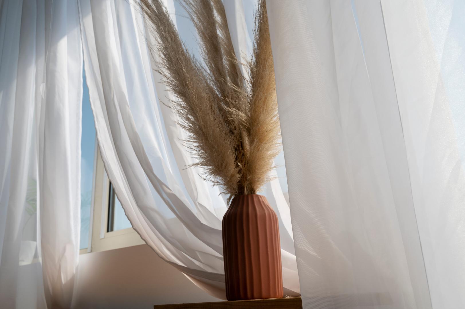 A windowsill with a beige vase and white polyester curtains.