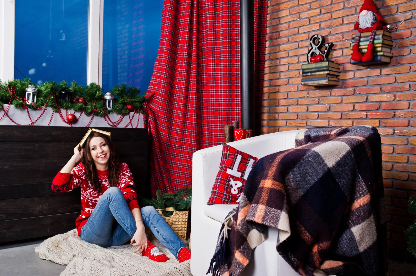 A girl wearing a Christmas sweater in a room decorated for Christmas with red plaid curtains.
