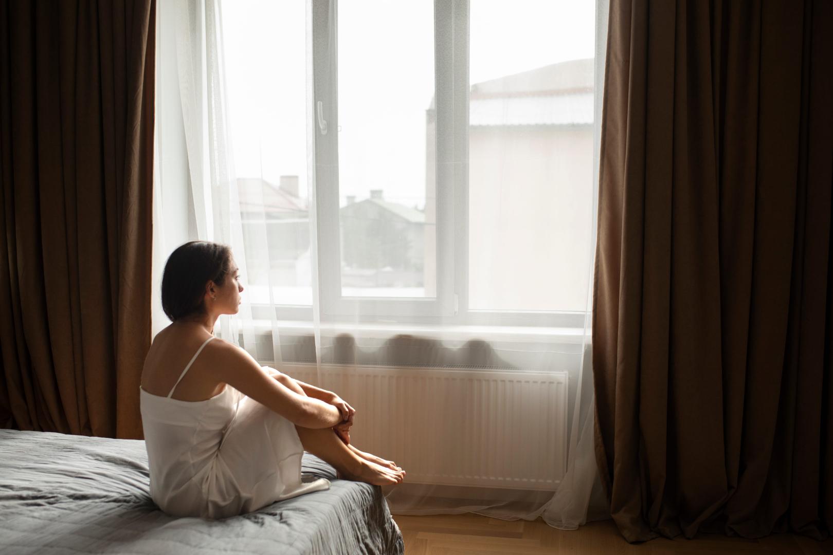 A woman sitting in a bedroom with white sheer curtains and brown blackout curtains.
