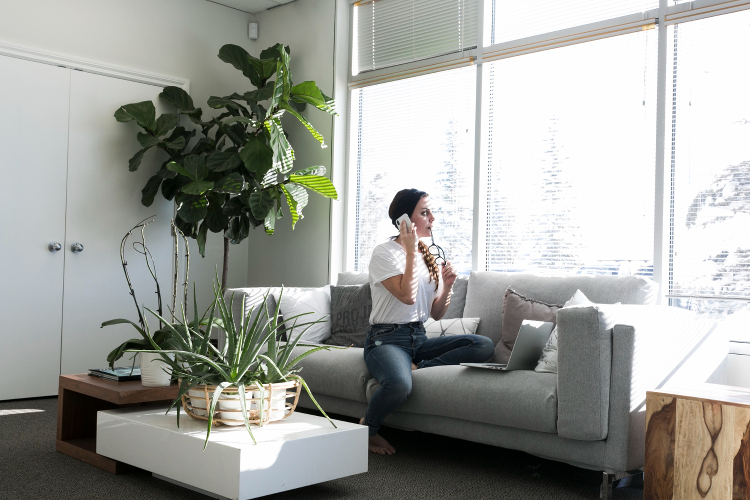 A woman sitting on a gray couch next to a large window with window blinds.