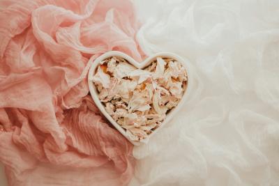 A heart-shaped white ceramic bowl filled with delicate, pale pink and cream dried flower petals. The bowl is centered on a backdrop of textured, gauzy fabric in two soft colors: dusty rose on the left and crisp white on the right. 