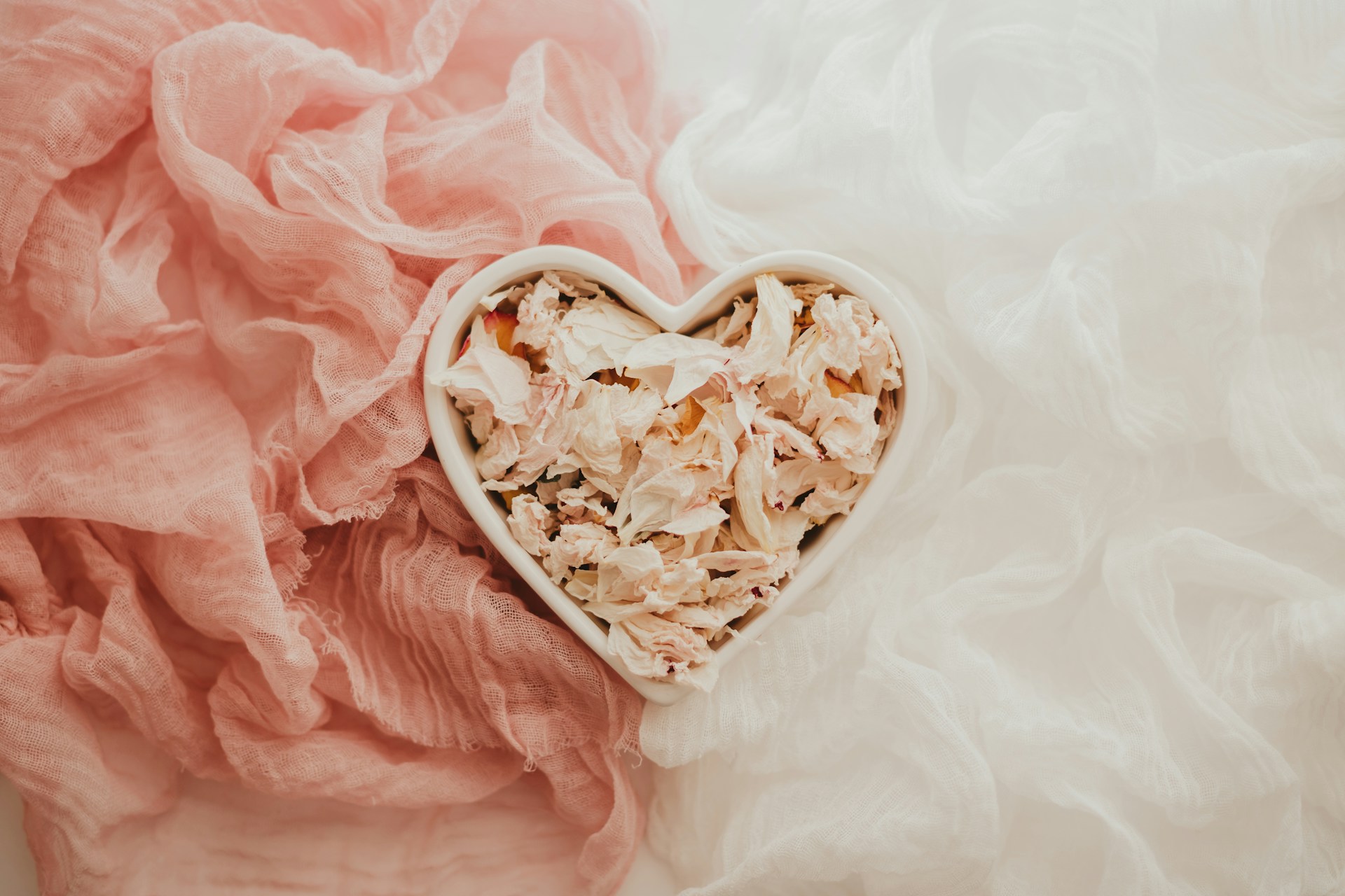 A heart-shaped white ceramic bowl filled with delicate, pale pink and cream dried flower petals. The bowl is centered on a backdrop of textured, gauzy fabric in two soft colors: dusty rose on the left and crisp white on the right. 