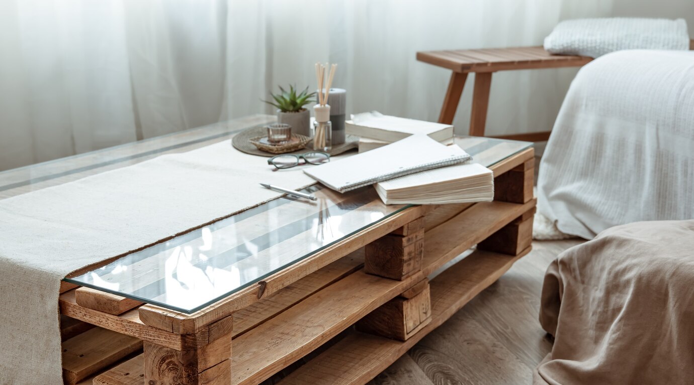 A table made out of wooden pallets and a sheet of glass next to the corner of a soft couch.