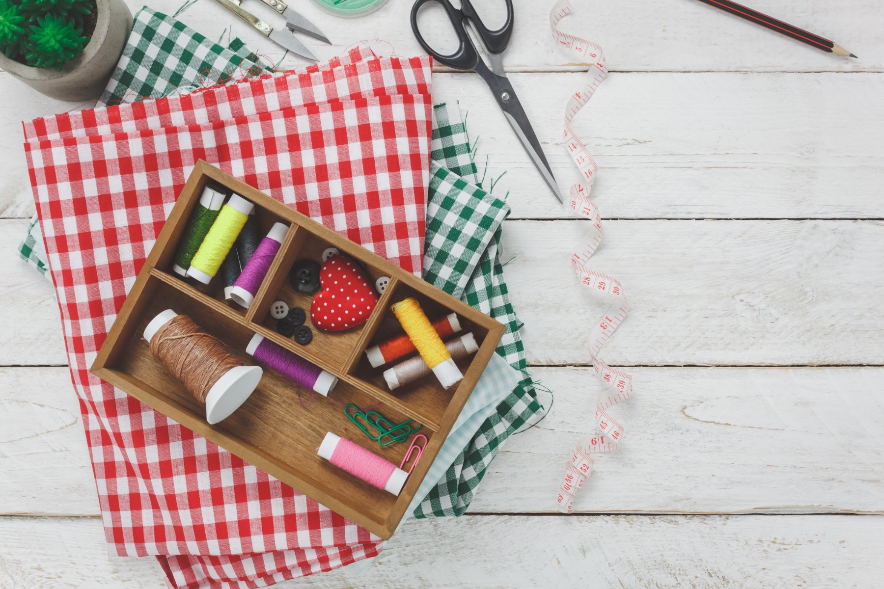 A wooden box filled with sewing supplies on top of green and red plaid fabrics, displayed on white wooden floor.