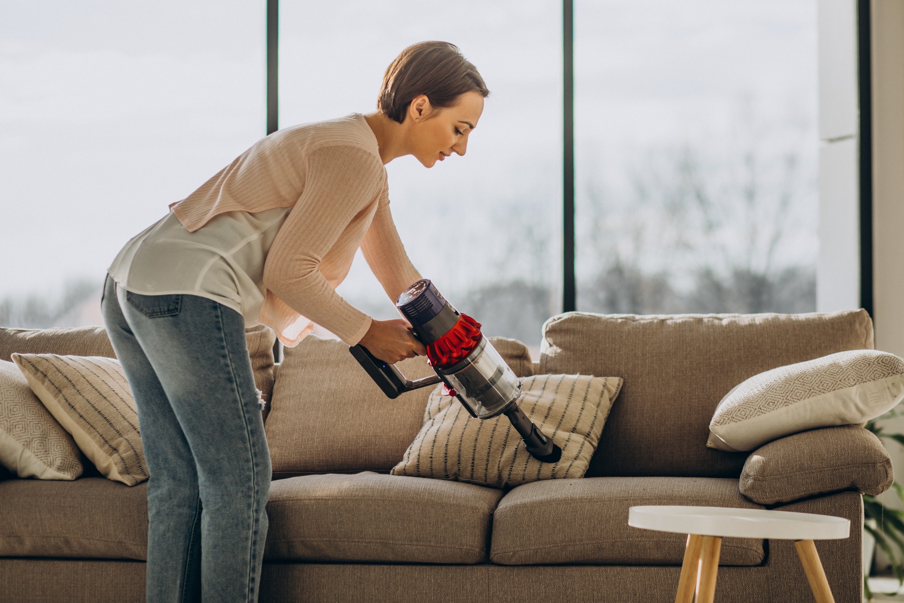 Young woman with rechargeable vacuum cleaner cleaning a couch.