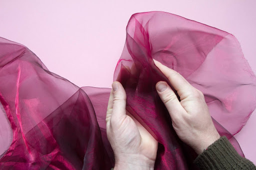 Hands holding a sheer burgundy organza fabric on a light pink background.